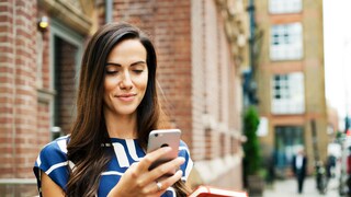 Frau in blau-weißem Shirt steht auf der Straße und schaut aufs Handy