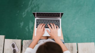 Frau sitzt auf Steg mit Füßen im Wasser und Laptop auf dem Schoß