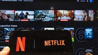 Smartphone mit großem Netflix-Logo auf dem Display. Im unscharfen Hintergrund sind verschiedene Film- und Seriencover zu sehen.
