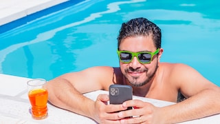 Mann steht im Pool, lehnt sich mit beiden Armen auf den Pool-Rand. Er trägt eine Sonnenbrille mit grünem Rahmen und hält ein Smartphone in der Hand. Neben ihm steht ein Glas mit einem orangem Getränk.