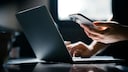Person vor einem Laptop mit einem Smartphone in der Hand.