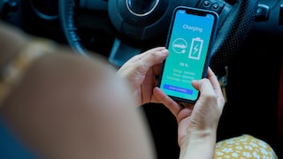 Person hält im Auto ein Smartphone mit Anzeige zum Ladevorgang eines Elektroautos in der Hand.