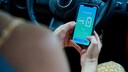 Person h&auml;lt im Auto ein Smartphone mit Anzeige zum Ladevorgang eines Elektroautos in der Hand.