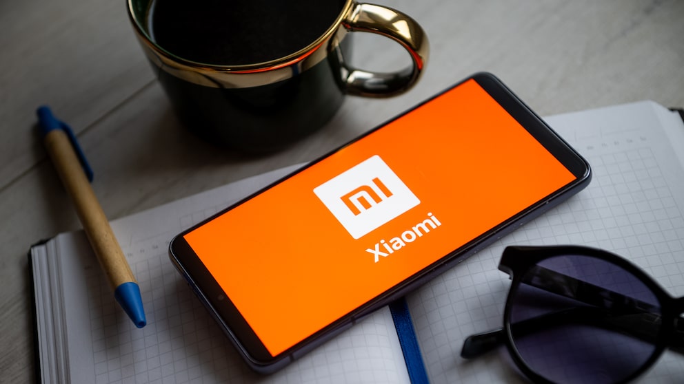 Ein Smartphone mit orangefarbenem Xiaomi-Startbildschirm liegt schräg auf einem karierten Notizbuch. Links daneben befindet sich ein Kugelschreiber mit blauem Griff. Oberhalb des Smartphones steht eine dunkle Tasse mit goldfarbenem Rand und Henkel. Rechts unten liegt eine dunkle Sonnenbrille teilweise auf dem Notizbuch. Alle Gegenstände befinden sich auf einer grauen Tischoberfläche.