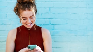 Frau vor türkisblauer Wand schaut auf ihr Smartphone und lächelt