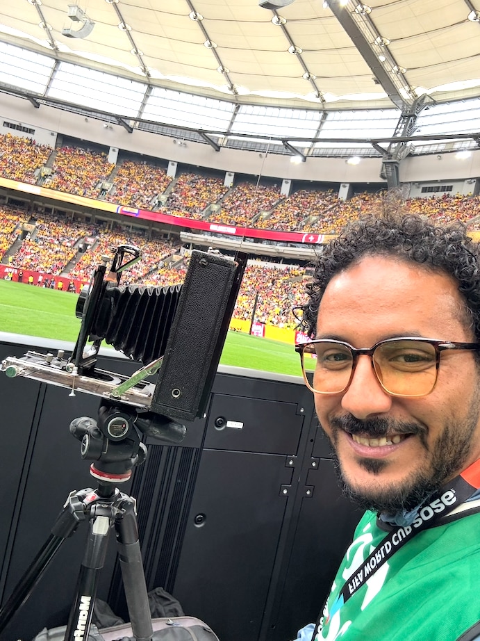 Selfie on the sidelines of a soccer stadium. In the foreground stands a historic large-format camera with bellows on a tripod. Next to it is a person with glasses and an accreditation band. In the background are the field, advertising boards, and crowded stands under the stadium's covered roof.
Provide your feedback on BizChat.
