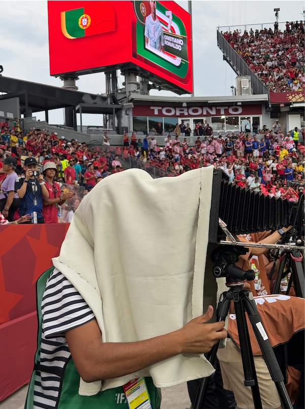 Person on the sidelines of a soccer stadium under a bright cloth in front of a historic large-format camera with bellows on a tripod. In the background are spectators in the stands and a large video screen with game graphics and the word 'TORONTO.' The photo was taken during a soccer match, with the camera aimed at the field.