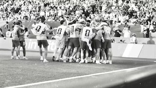 Schwarz-Weiß-Fotografie einer jubelnden Fußballmannschaft an der Seitenlinie eines Stadions. Mehrere Spieler und Ersatzspieler haben sich zu einer Traube zusammengeschlossen und feiern vor den Werbebanden am Spielfeldrand. Im Hintergrund sind voll besetzte Tribünen mit zahlreichen Zuschauern zu sehen.