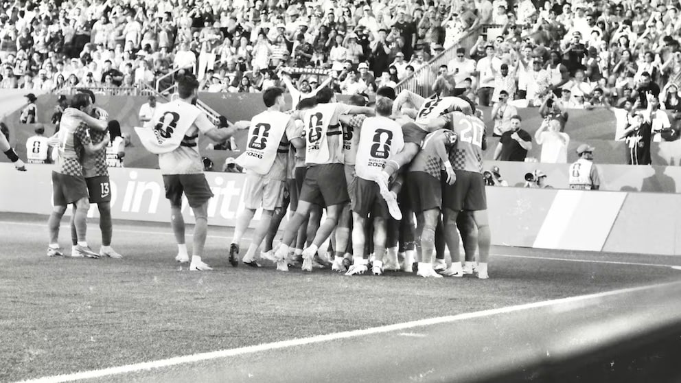 Schwarz-Weiß-Fotografie einer jubelnden Fußballmannschaft an der Seitenlinie eines Stadions. Mehrere Spieler und Ersatzspieler haben sich zu einer Traube zusammengeschlossen und feiern vor den Werbebanden am Spielfeldrand. Im Hintergrund sind voll besetzte Tribünen mit zahlreichen Zuschauern zu sehen.