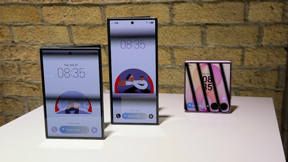 Drei Samsung-Falt-Smartphones stehen auf einem weißen Tisch vor einer hellen Steinwand. Links steht ein aufgeklapptes Gerät im Querformat mit großem Display, in der Mitte ein aufgeklapptes Gerät im Hochformat mit schmaleren Bildschirmrändern, rechts ein teilweise zusammengeklapptes Smartphone in Rosa. Auf allen Bildschirmen ist die Uhrzeit 08:35 zu sehen. Die beiden größeren Geräte zeigen zusätzlich eine Illustration auf dem Sperrbildschirm. Die Aufnahme entstand in einem Innenraum mit warmem Licht.