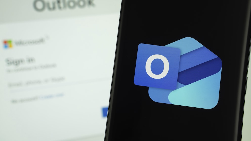 Outlook-Logo auf dem Smartphone, im Hintergrund Outlook-Anmeldefenster auf einem Computer