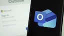 Outlook-Logo auf dem Smartphone, im Hintergrund Outlook-Anmeldefenster auf einem Computer