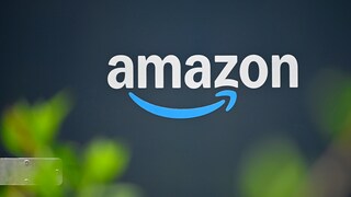Amazon-Logo in weißer Schrift auf dunkelblauem Hintergrund. Unter dem Schriftzug befindet sich der blaue Pfeil des Unternehmens. Im Vordergrund sind unscharfe grüne Pflanzen zu sehen.