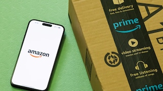 Ein Smartphone mit geöffnetem Amazon-Logo liegt auf einer grünen Oberfläche neben einem Amazon-Paket. Auf dem Paket ist schwarzes Prime-Klebeband mit Symbolen und englischen Hinweisen zu Versand, Video-Streaming und Musik zu sehen.