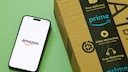 Ein Smartphone mit ge&ouml;ffnetem Amazon-Logo liegt auf einer gr&uuml;nen Oberfl&auml;che neben einem Amazon-Paket. Auf dem Paket ist schwarzes Prime-Klebeband mit Symbolen und englischen Hinweisen zu Versand, Video-Streaming und Musik zu sehen.