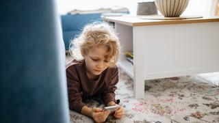 Kind liegt auf einem Teppich und schaut auf ein Smartphone in einem Wohnzimmer.