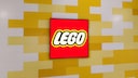 Rotes Lego-Logo auf einem rechteckigen Bildschirm vor einer Wand aus versetzt angeordneten gelben und wei&szlig;en rechteckigen Elementen. Das Logo befindet sich mittig im Bild und ist von einem schwarzen Rahmen umgeben. Die Wand im Hintergrund erinnert mit ihrer Gestaltung an gestapelte Lego-Steine.