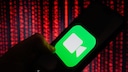 Eine Hand h&auml;lt ein Smartphone mit gro&szlig; dargestelltem FaceTime-Symbol auf dem Display. Im Hintergrund sind unscharfe rote Lichtmuster zu sehen, die an digitale Datenstr&ouml;me erinnern.