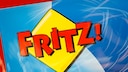 Das Bild zeigt das rote Fritz!-Logo auf gelbem, rautenf&ouml;rmigem Hintergrund. Dahinter sind blaue und hellblaue Fl&auml;chen mit wei&szlig;en Linien zu sehen.