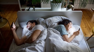 Zwei Personen liegen in einem Doppelbett mit dem Rücken zueinander und schauen jeweils auf ein Smartphone. Das Bett steht in einem hellen Schlafzimmer mit Holzboden, Zimmerpflanzen und einer Nachttischlampe. Das Licht der Smartphone-Displays beleuchtet ihre Gesichter.
