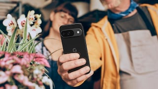 Eine Person hält ein schwarzes Google-Pixel-Smartphone in die Kamera. Das Gerät ist scharf im Vordergrund zu sehen, die Rückseite mit dem Google-Logo und dem horizontalen Kameramodul ist deutlich erkennbar. Im Hintergrund befinden sich zwei unscharf dargestellte Personen sowie mehrere Blumen und Pflanzen. Die Aufnahme entstand offenbar in einem Innenraum mit warmer Beleuchtung.