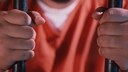 Nahaufnahme von zwei H&auml;nden, die Gitterst&auml;be festhalten. Im Hintergrund ist eine Person in orangefarbener Kleidung unscharf zu sehen. Das Bild symbolisiert eine Gef&auml;ngnissituation.