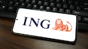 Ein Smartphone liegt waagerecht auf einem dunklen Holztisch vor einer schwarzen Tastatur. Auf dem hellen Bildschirm sind das blaue ING-Logo und das orangefarbene L&ouml;wen-Symbol der Bank zu sehen. Das Smartphone ist leicht schr&auml;g ausgerichtet und f&uuml;llt den Gro&szlig;teil des Bildes aus.