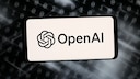 Ein Smartphone oder Display zeigt auf hellem Hintergrund das OpenAI-Logo und den Schriftzug &bdquo;OpenAI&ldquo;. Im Hintergrund sind unscharfe Reihen aus Nullen und Einsen sowie helle Lichtstreifen auf dunklem Grund zu sehen. Die Darstellung wirkt wie eine grafische Illustration zu den Themen K&uuml;nstliche Intelligenz, Software und Cybersicherheit.