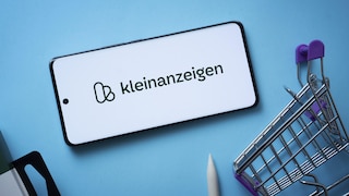 Ein Smartphone liegt waagerecht auf einem hellblauen Hintergrund. Auf dem Display ist das Logo und der Schriftzug „kleinanzeigen“ zu sehen. Rechts neben dem Smartphone steht ein kleiner Metall-Einkaufswagen mit violetten Kunststoffteilen. Am unteren Bildrand sind zudem die Spitze eines weißen Stifts und ein dunkler Gegenstand teilweise sichtbar.