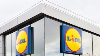 Außenansicht einer Lidl-Filiale mit zwei großen blau-gelben Lidl-Schildern an einer Glasfassade. Das Gebäude hat helle Fassadenelemente, große Fenster und ein flaches Dach mit Geländer.