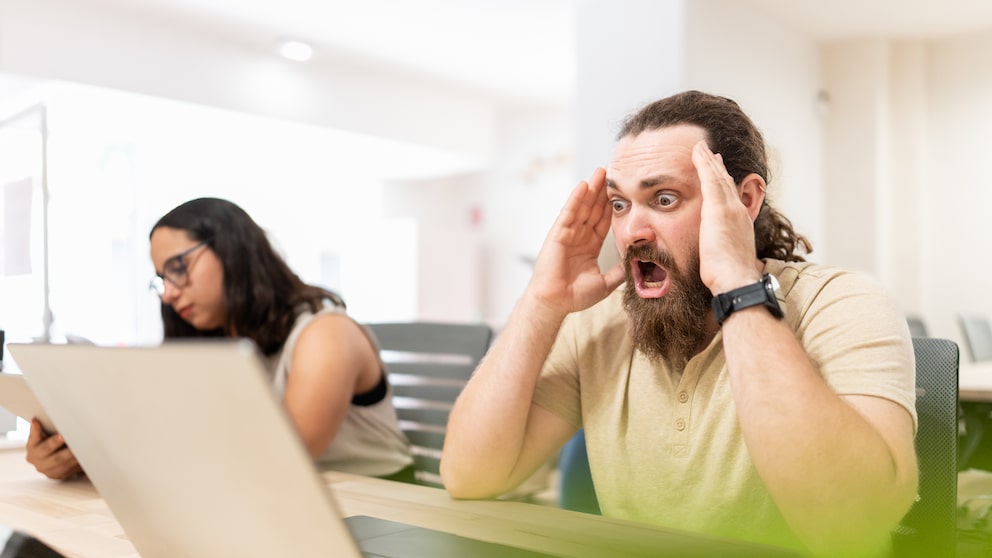 Eine Person sitzt in einem hellen Büro vor einem Laptop und hält beide Hände an den Kopf. Im Hintergrund sitzt eine weitere Person an einem Laptop.