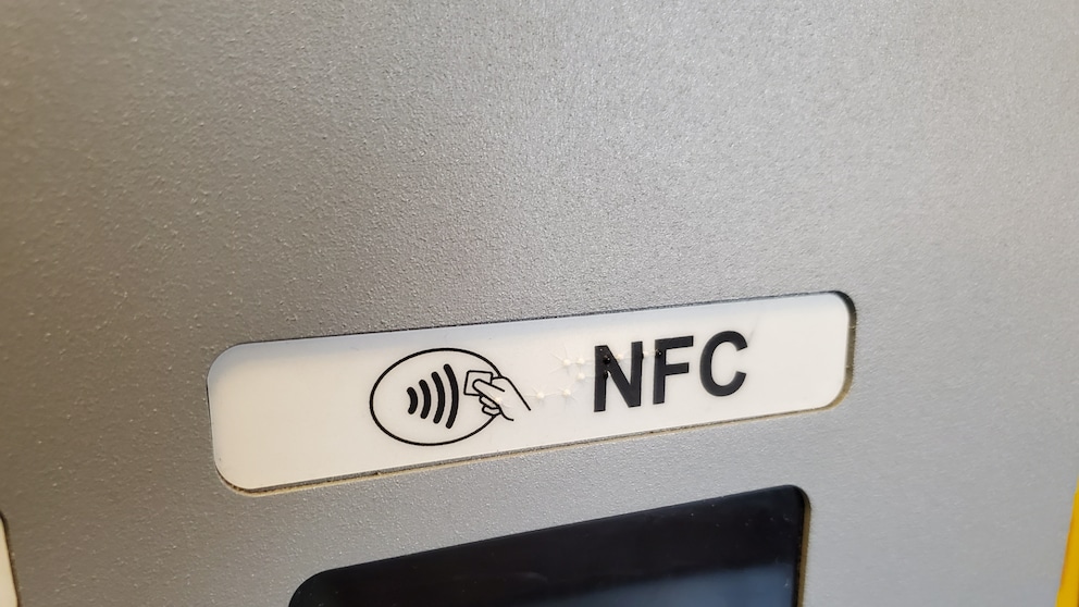 Nahaufnahme eines NFC-Symbols mit der Aufschrift „NFC“ an einem Kartenlesegerät oder Geldautomaten. Unterhalb des Symbols ist ein Teil des Displays zu sehen.