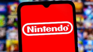Ein Smartphone mit dem Nintendo-Logo auf rotem Bildschirm steht im Vordergrund. Im unscharfen Hintergrund sind zahlreiche bunte Spiele-Cover zu sehen.