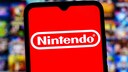 Ein Smartphone mit dem Nintendo-Logo auf rotem Bildschirm steht im Vordergrund. Im unscharfen Hintergrund sind zahlreiche bunte Spiele-Cover zu sehen.