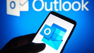 Ein Smartphone wird vor einem blau beleuchteten Hintergrund mit Outlook-Logo gehalten. Auf dem Display ist das Symbol der Outlook-App mit blauem Briefumschlag und weißem „O“ zu sehen. Im Hintergrund ist der Schriftzug „Outlook“ unscharf dargestellt.