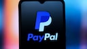 Smartphone mit schwarzem Display, auf dem das blaue und wei&szlig;e PayPal-Logo sowie der Schriftzug &bdquo;PayPal&ldquo; zu sehen sind. Der Hintergrund ist unscharf und zeigt einen Farbverlauf in Blau-, Rosa- und Oranget&ouml;nen.
