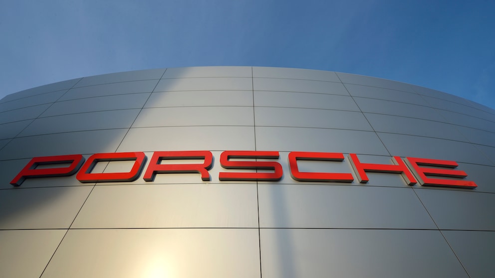 Porsche-Schriftzug in roten Buchstaben an einer silberfarbenen Gebäudefassade, aufgenommen aus niedriger Perspektive vor blauem Himmel.