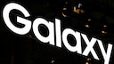Leuchtender Samsung-Galaxy-Schriftzug vor dunklem Hintergrund. Zu sehen sind vor allem die gro&szlig;en wei&szlig;en Buchstaben &bdquo;Galaxy&ldquo;, die schr&auml;g durch das Bild laufen. Im Hintergrund sind nur wenige dunkle Strukturen und kleine Lichtpunkte erkennbar.