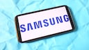 Ein Smartphone liegt waagerecht auf einem hellblauen, zerknitterten Untergrund. Auf dem Display ist auf wei&szlig;em Hintergrund das blaue Samsung-Logo in gro&szlig;en Gro&szlig;buchstaben zu sehen. Das Ger&auml;t hat einen dunklen Rahmen mit leicht bronzefarbener Kante. Das Smartphone ist schr&auml;g im Bild platziert und nimmt den Gro&szlig;teil der Aufnahme ein.