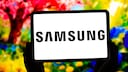 Ein Tablet wird mit beiden H&auml;nden vor einem unscharfen, bunten Hintergrund gehalten. Auf dem wei&szlig;en Display ist mittig das schwarze Samsung-Logo zu sehen. Im Hintergrund sind kr&auml;ftige Rot-, Gelb-, Gr&uuml;n- und Blaut&ouml;ne erkennbar.