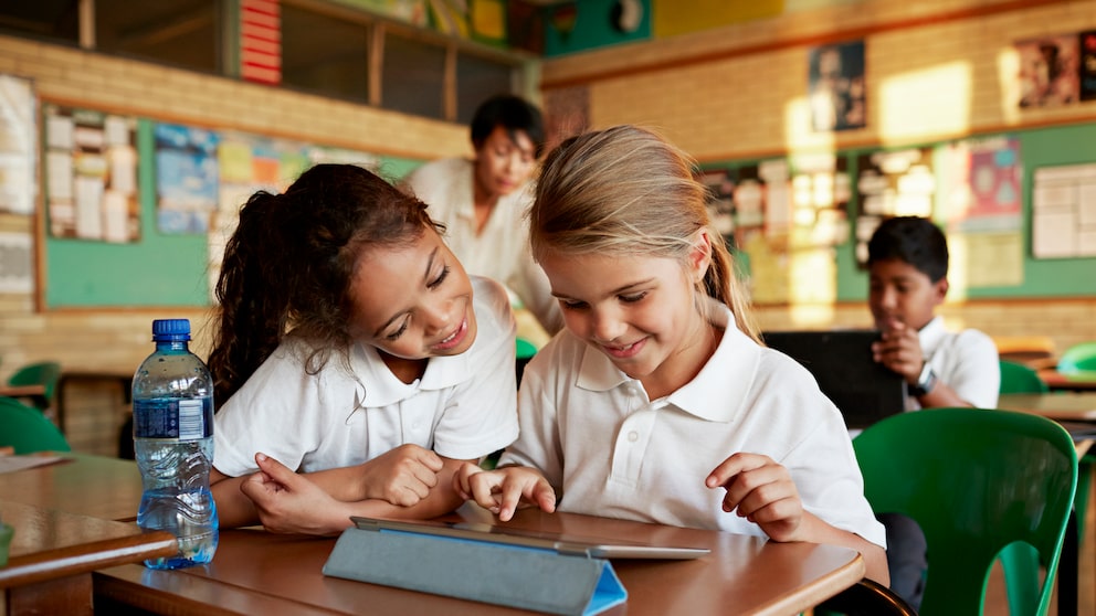 Zwei Grundschulkinder sitzen gemeinsam an einem Tisch in einem Klassenzimmer und schauen auf ein Tablet. Im Hintergrund sind weitere Kinder sowie eine Lehrkraft zu sehen. Auf dem Tisch steht außerdem eine Wasserflasche.