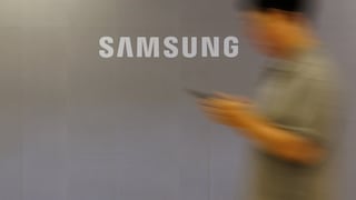 Großes Samsung-Logo auf einer hellgrauen Wand. Rechts im Bild ist eine unscharf dargestellte Person zu sehen, die ein Smartphone in der Hand hält und sich an der Wand vorbeibewegt. Die Aufnahme ist bewusst mit Bewegungsunschärfe gestaltet, wodurch das weiße Samsung-Logo im Mittelpunkt steht.
