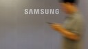 Gro&szlig;es Samsung-Logo auf einer hellgrauen Wand. Rechts im Bild ist eine unscharf dargestellte Person zu sehen, die ein Smartphone in der Hand h&auml;lt und sich an der Wand vorbeibewegt. Die Aufnahme ist bewusst mit Bewegungsunsch&auml;rfe gestaltet, wodurch das wei&szlig;e Samsung-Logo im Mittelpunkt steht.