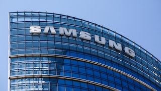 Großaufnahme der oberen Stockwerke eines modernen Bürogebäudes mit gebogener Glasfassade. Auf der Fassade sind große weiße Buchstaben mit dem Schriftzug „SAMSUNG“ angebracht. Der Himmel im Hintergrund ist klar und blau.