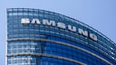Gro&szlig;aufnahme der oberen Stockwerke eines modernen B&uuml;rogeb&auml;udes mit gebogener Glasfassade. Auf der Fassade sind gro&szlig;e wei&szlig;e Buchstaben mit dem Schriftzug &bdquo;SAMSUNG&ldquo; angebracht. Der Himmel im Hintergrund ist klar und blau.