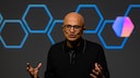 Satya Nadella mit Brille und dunklem Oberteil steht auf einer B&uuml;hne und h&auml;lt beide H&auml;nde leicht erhoben. Im Hintergrund sind mehrere blaue sechseckige Liniengrafiken sowie ein unscharfes sechseckiges Symbol in Blau und Magenta auf dunklem Hintergrund zu sehen.