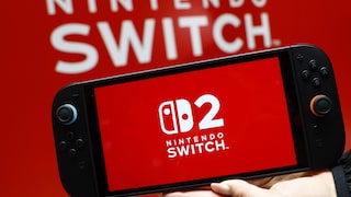 Eine Person hält eine Nintendo Switch 2 mit eingeschaltetem Startbildschirm vor einem roten Hintergrund mit dem Nintendo-Switch-Logo. Die Konsole ist frontal zu sehen und wird mit einer Hand von unten gestützt.
