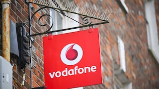 Ein rotes Vodafone-Schild mit weißem Logo und Schriftzug hängt an einer schwarzen Metallhalterung an einer Backsteinfassade. Oberhalb der Halterung sind Taubenabwehrspitzen angebracht, im Hintergrund sind weitere Teile des Gebäudes unscharf zu sehen.