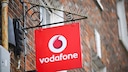 Ein rotes Vodafone-Schild mit wei&szlig;em Logo und Schriftzug h&auml;ngt an einer schwarzen Metallhalterung an einer Backsteinfassade. Oberhalb der Halterung sind Taubenabwehrspitzen angebracht, im Hintergrund sind weitere Teile des Geb&auml;udes unscharf zu sehen.