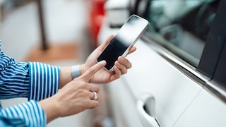 Eine Person hält ein iPhone vor die Fahrertür eines weißen Autos und tippt auf den Bildschirm. Das Smartphone befindet sich direkt neben dem Türgriff, im Hintergrund sind weitere Fahrzeuge unscharf zu sehen.